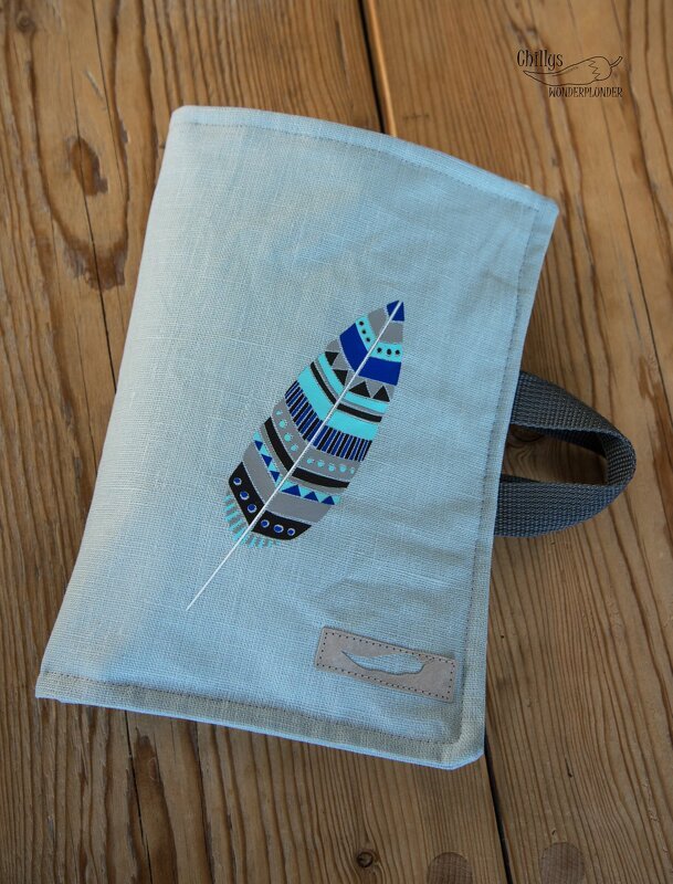 Heller Leinenstoff mit großem grafischem Federmotiv in Blau, Türkis, Grau und Schwarz sowie einem kleinen Lederetikett auf Holzuntergrund.