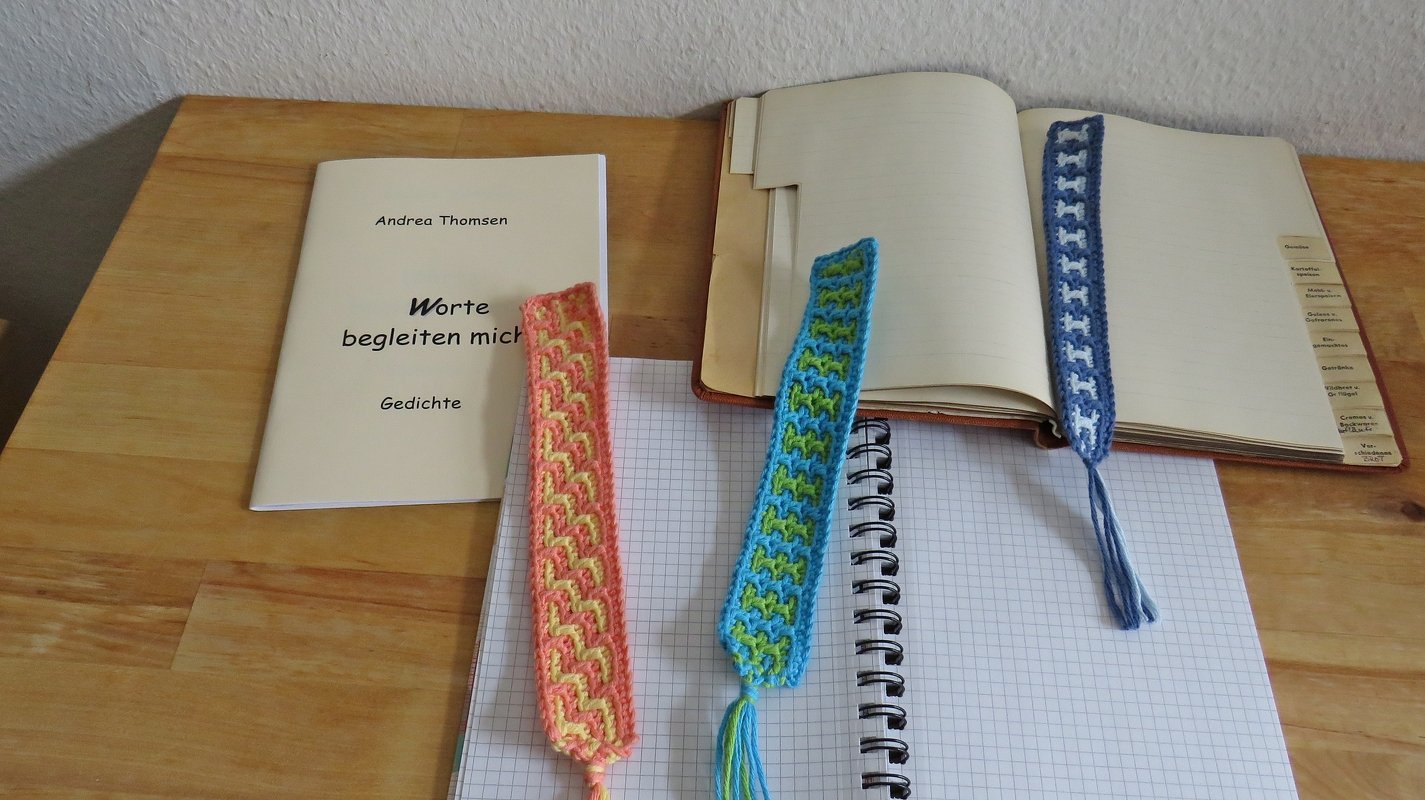 Drei gehäkelte Lesezeichen auf einer Holzoberfläche: ein blau-weißes mit wiederkehrendem Motiv in einem offenen Buch und zwei bunte geometrische Lesezeichen auf einem Notizbuch.