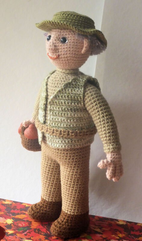 Stehende Amigurumi-Puppe mit gehäkeltem Hut, Weste und Korb mit orangefarbenen Fundstücken.
