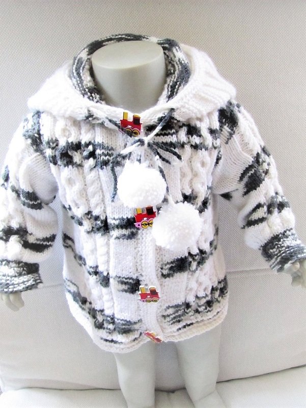Gestrickte Babyjacke in Weiß und Grau mit Kapuze, Zopfmuster, Knöpfen mit Zug‑Motiv und Pompons an einer Kinderpuppe.