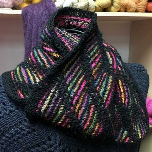 Black knitted cowl with bright pink, yellow, orange, green, and blue diagonal stripes on a shelf