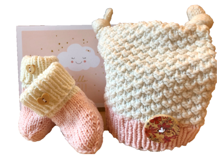 Gestrickte Babymütze in Creme mit rosa Rand und großem Dekoknopf, daneben rosa-creme Babyschühchen mit kleinen Herzknöpfen