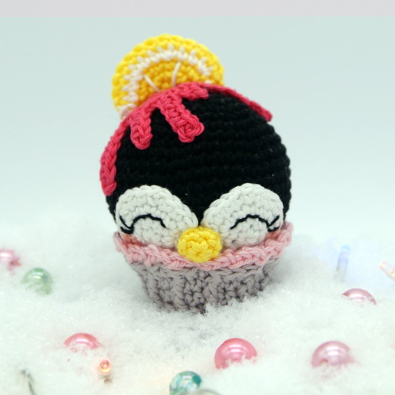 Gehäkelte Pinguin-Cupcake-Figur in Schwarz, Weiß und Gelb mit rosa Topping auf künstlichem Schnee, umgeben von pastellfarbenen Kugeln.