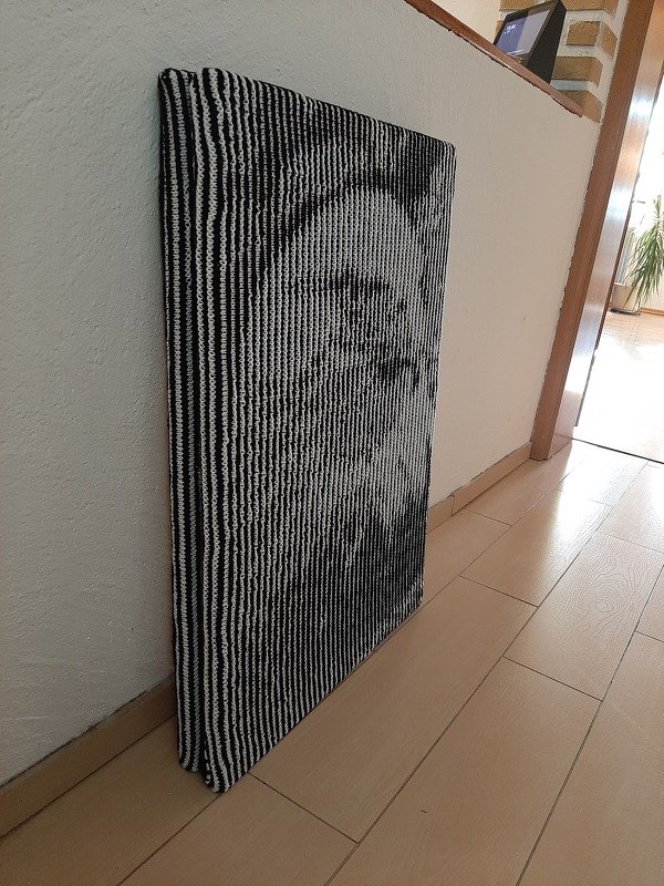 Gestricktes schwarz-weißes Wandbild mit stilisiertem Gesicht, auf Rahmen gespannt und an Wand gelehnt