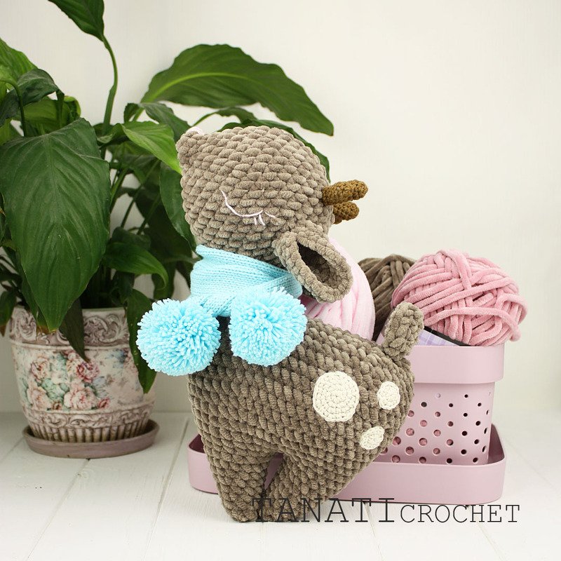 Gehäkeltes braunes Amigurumi-Reh mit hellblauem Schal und Bommeln, neben Garnknäueln und einer Zimmerpflanze.