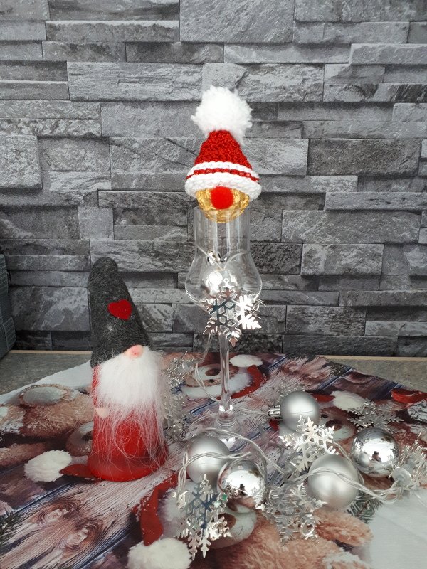 Rot-weiße gehäkelte Mini-Weihnachtsmütze mit Bommel auf umgedrehtem Weinglas, umgeben von Kugeln und Wichtel.