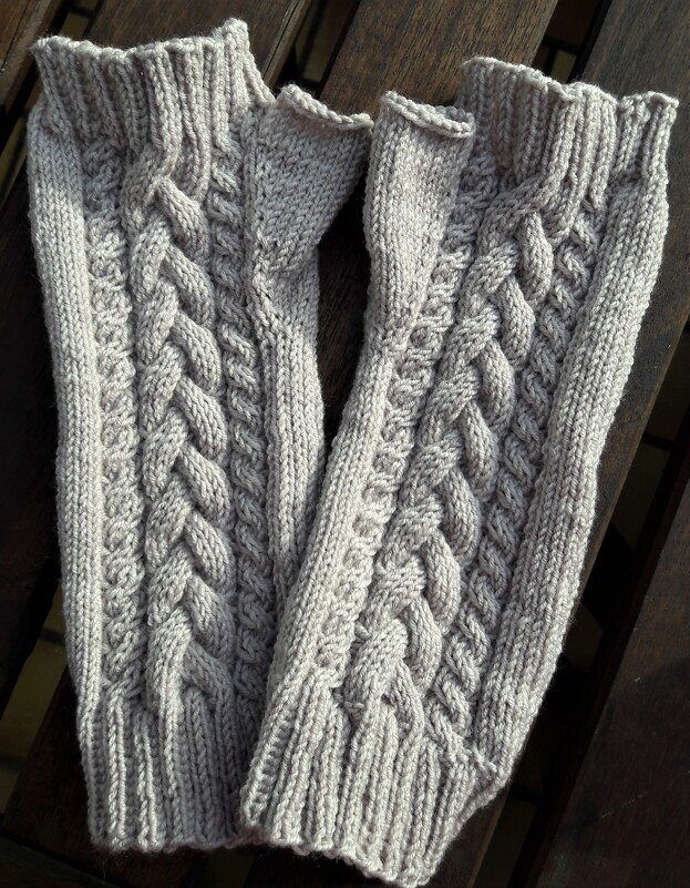 Ein Paar beigefarbene, gestrickte Armstulpen mit Zopfmuster auf einer Holzoberfläche.