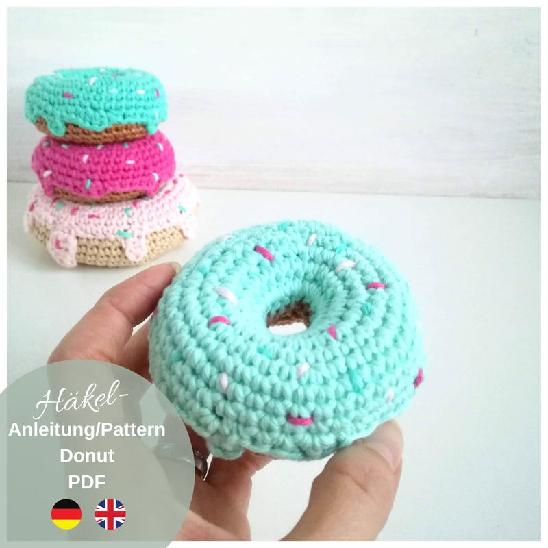 Hand holding a mint-green crocheted donut toy with embroidered sprinkles and stacked pastel crocheted donuts in the background