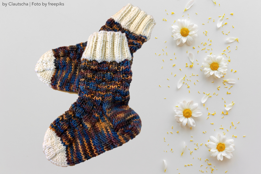 Gestrickte Kindersocken aus meliertem Garn mit cremefarbenen Zehen und Rippbündchen auf hellem Hintergrund neben Gänseblümchen