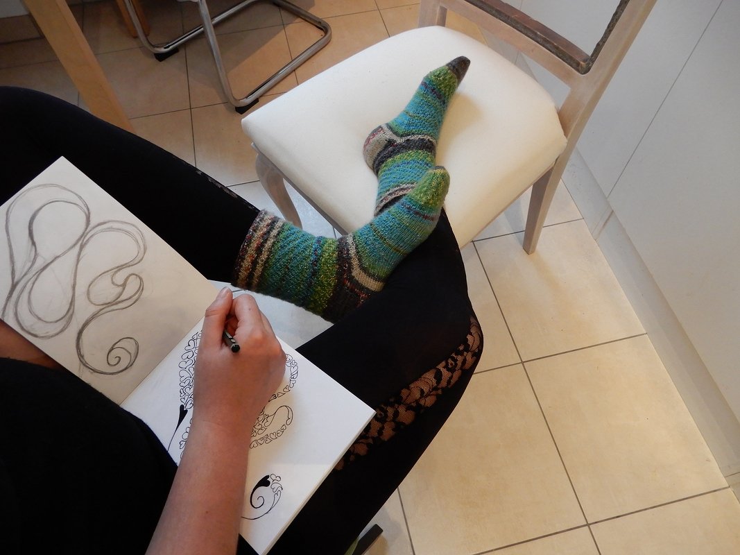Seated person wearing green-blue striped knitted socks and sketching in a notebook