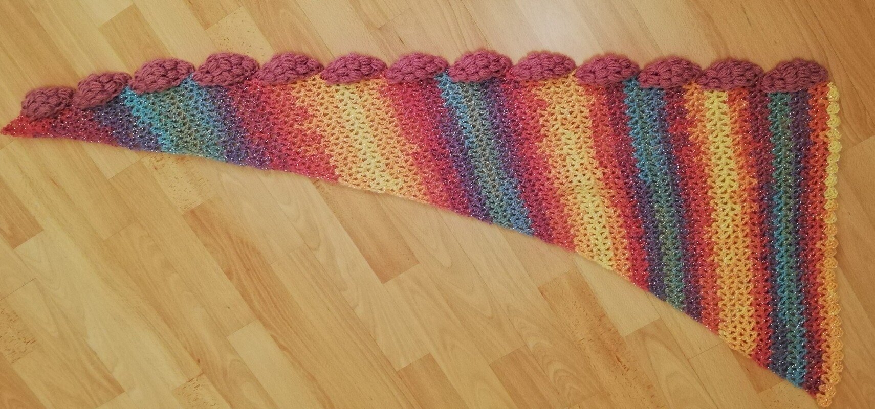 Gehäkelter Drachenschwanz-Schal, Regenbogen-Farbverlauf, Glitzereffekt und rosa Noppenkante auf Holzboden