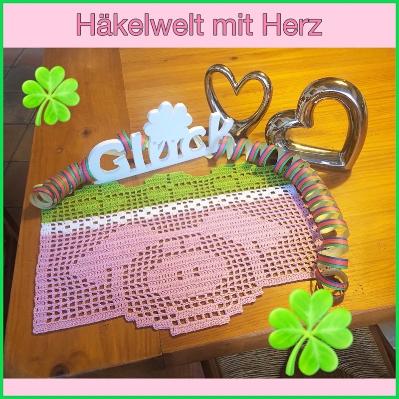 Gehäkeltes rosa Deckchen mit Herzmuster und grünem Rand auf Holztisch, daneben Dekoherzen und Schriftzug 'Glück'.