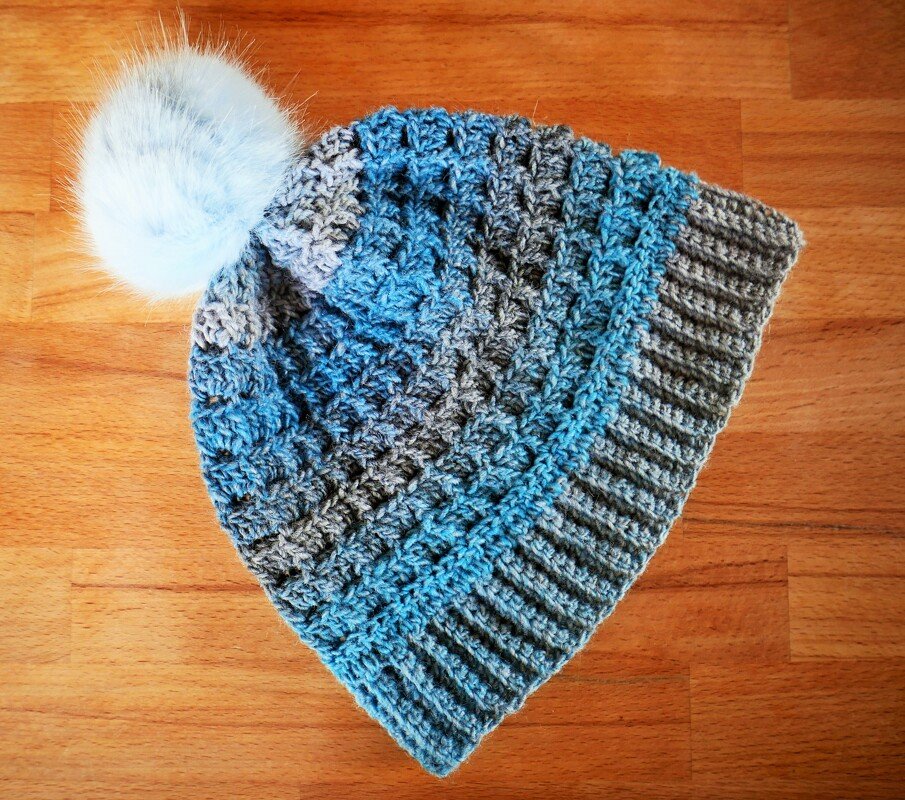 Gehäkelte blau-graue Damenmütze mit Rippbündchen und weißem Bommel auf Holzuntergrund