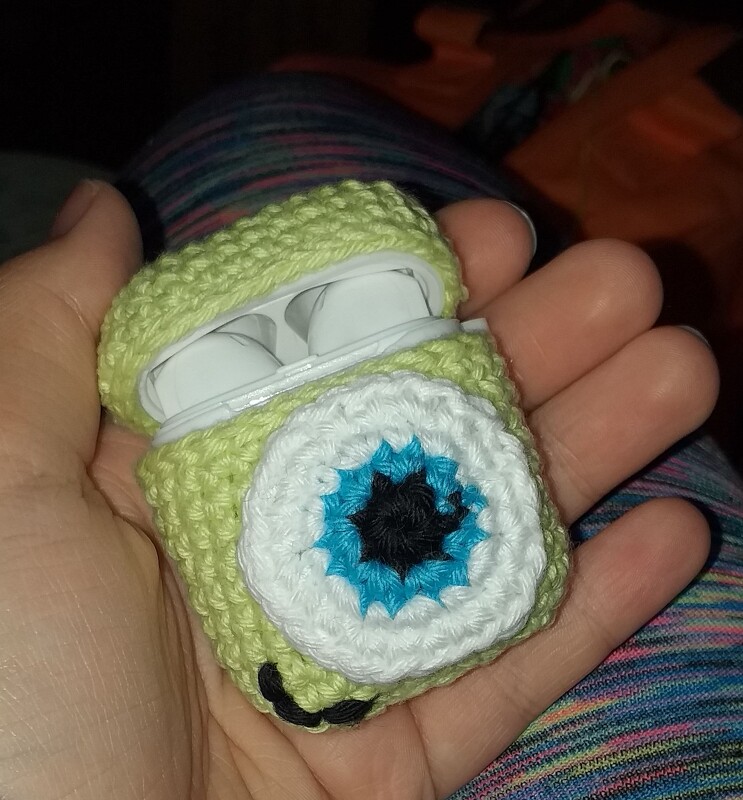 Geöffnete, handgehäkelte hellgrüne Hülle mit Monstergesicht (Auge und Mund) hält weiße kabellose Ohrhörer in einer Hand.