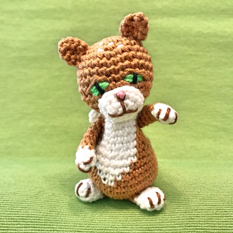 Braun-weißes gehäkeltes Amigurumi-Kätzchen mit grünen, gestickten Augen auf grünem Hintergrund