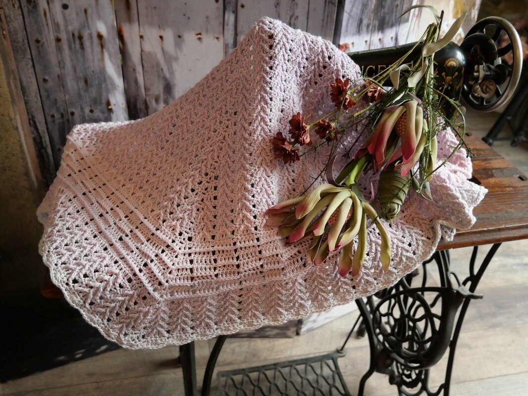 Helllila gehäkeltes Dreieckstuch mit Spitzenmuster und getrockneten Blumen auf Holztisch mit Nähmaschinengestell.