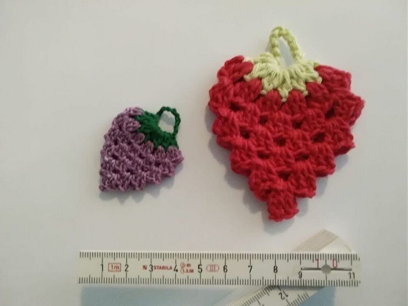 Zwei kleine gehäkelte Früchte (Erdbeere und Traube) auf hellem Hintergrund, mit Lineal zur Größenreferenz.