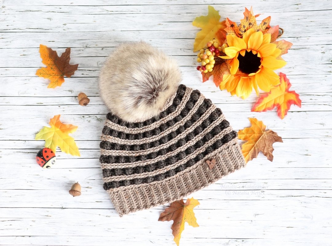 Braun-beige gehäkelte Bommelmütze mit Noppenmuster und Herzanhänger auf der Krempe, umgeben von Herbstblättern und Sonnenblume auf hellem Holz.