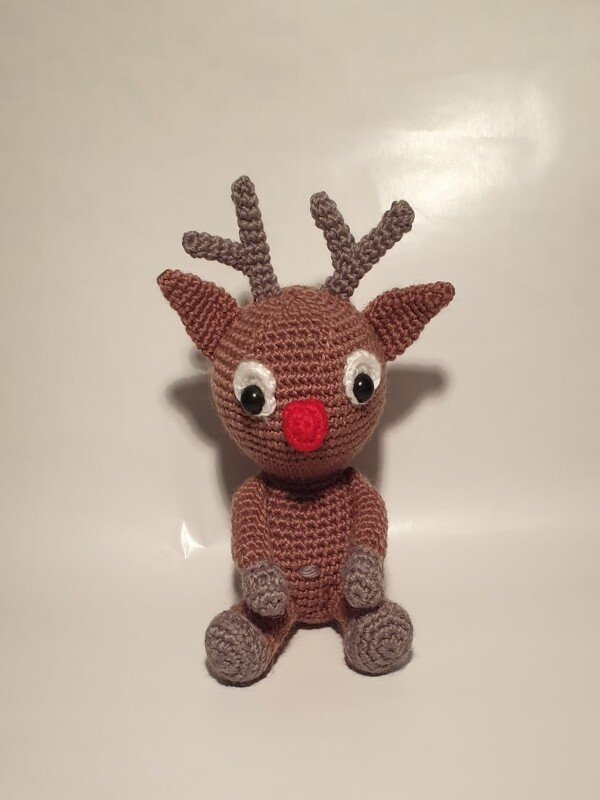 Sitzendes braunes Amigurumi-Rentier mit roter Nase und grauem Geweih vor hellem Hintergrund.