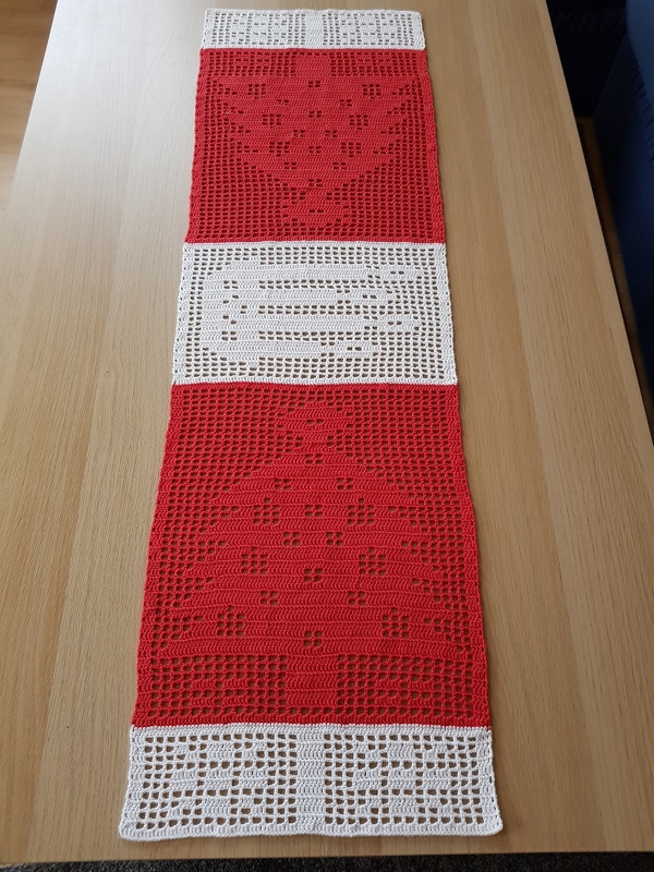 Red and white filet crochet table runner spelling "NOEL" with patterned motifs, displayed on a wooden surface.
