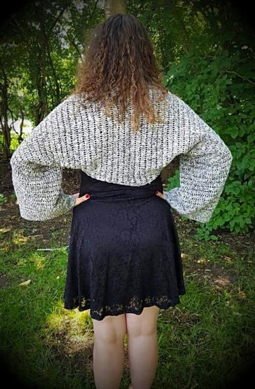 Rückenansicht einer Person mit grau-meliertem, gehäkeltem Shrug mit weiten Ärmeln über schwarzem Kleid vor grünem Hintergrund.