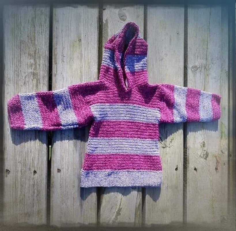 Gehäkelter Kinder-Kapuzenpullover in gestreiften Pink- und Lilatönen, flach auf Holzbrettern gelegt