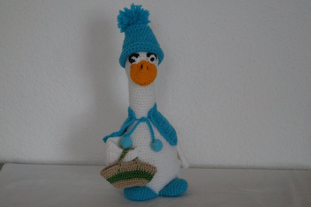 Weiß gehäkelte Amigurumi-Gans mit orangefarbenem Schnabel, türkisfarbener Mütze, Schal und Korb