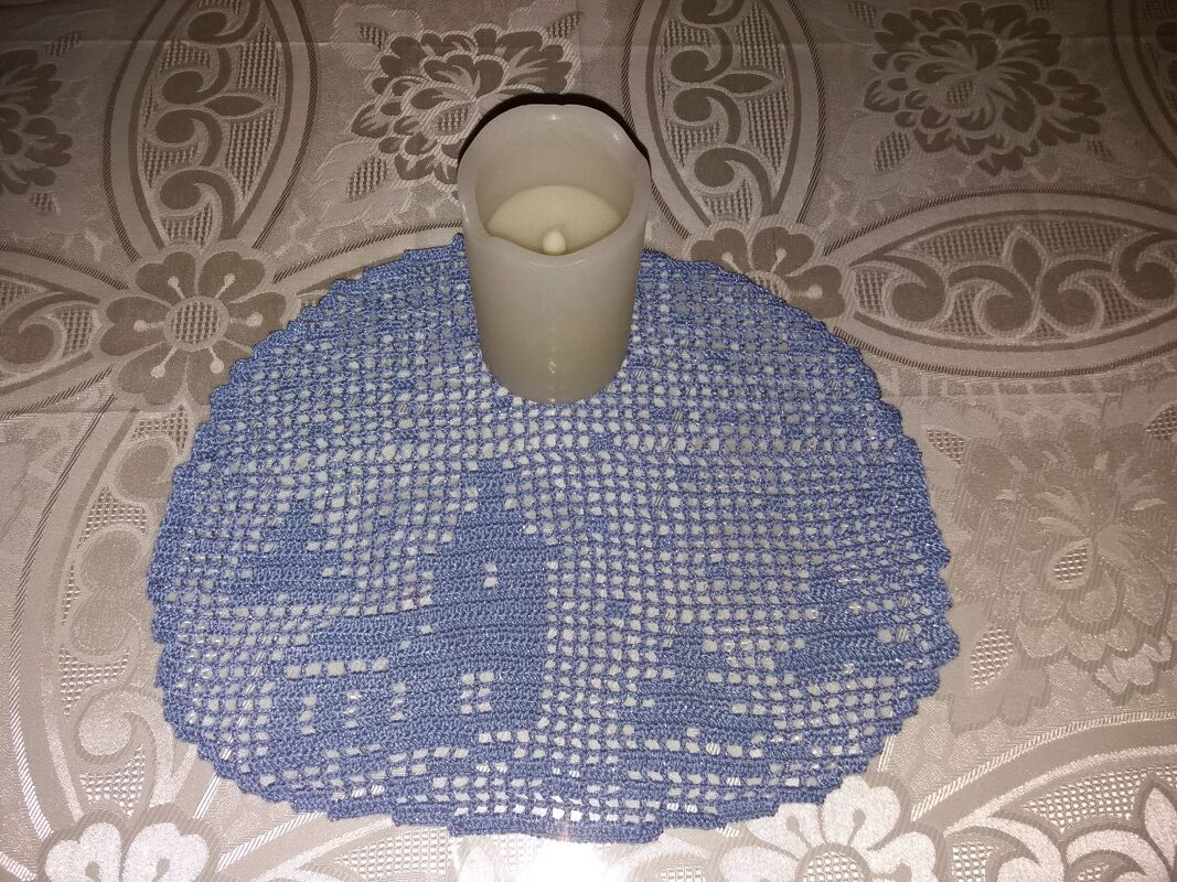 Rundes hellblaues gehäkeltes Filet-Deckchen auf gemustertem Tischtuch mit einer Kerze