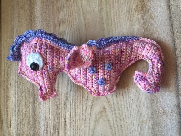 Rosa-lila gehäkeltes Seepferdchen mit Wackelaugen auf heller Holzoberfläche.