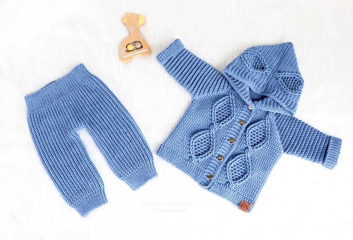 Light blue crocheted baby set with a hooded cardigan decorated with leaf motifs and matching ribbed trousers on a white faux-fur surface next to a wooden giraffe rattle.