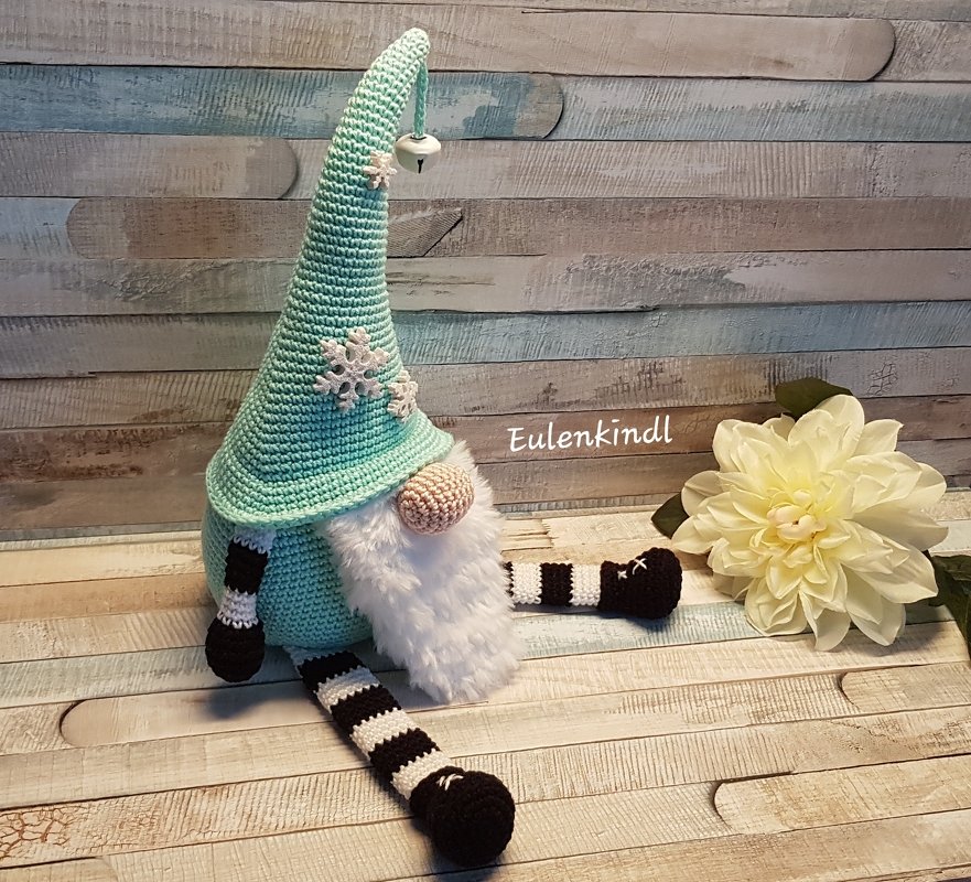 Sitzender, gehäkelter Wichtel mit mintgrüner Zipfelmütze, weißem Bart, beiger Nase und schwarz-weiß gestreiften Beinen, dekoriert mit Schneeflocken und kleinem Glöckchen.