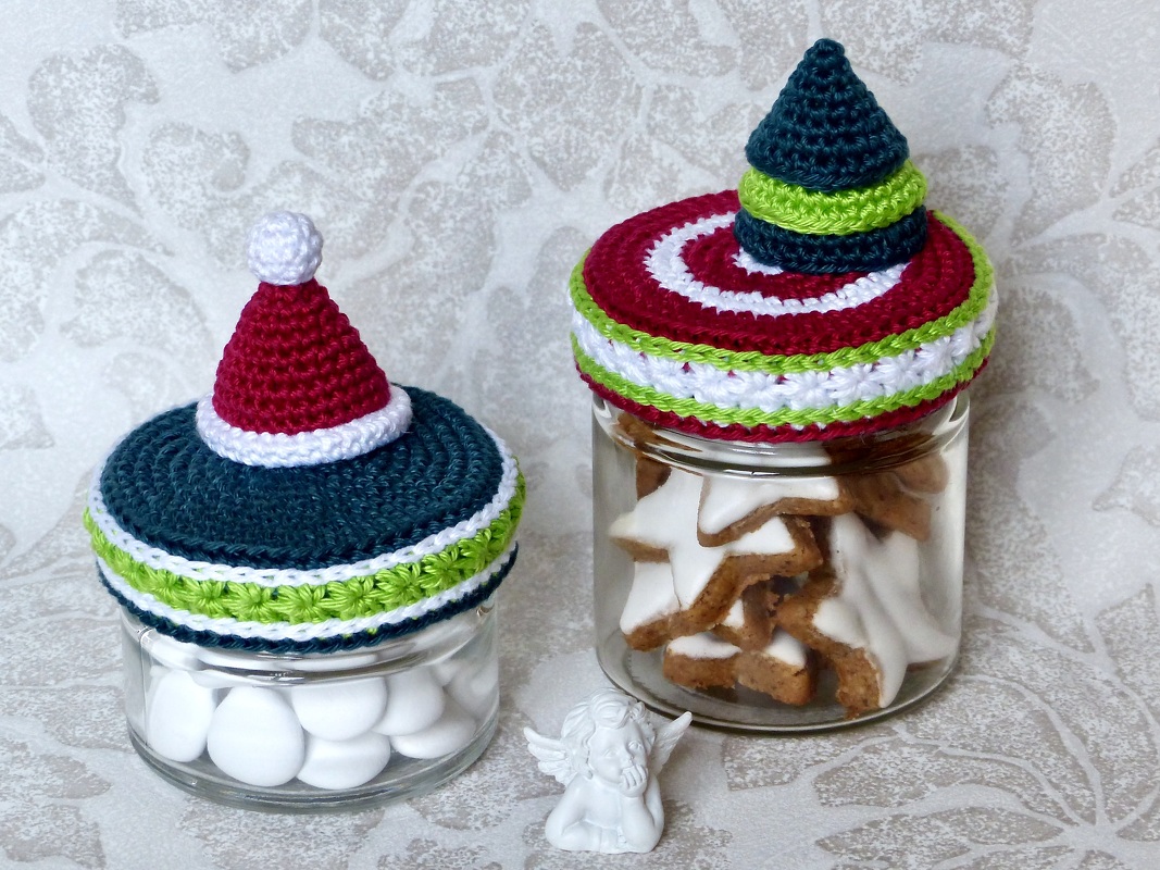 Zwei Glasbehälter mit handgehäkelten Weihnachtsdeckeln, gefüllt mit Sternkeksen und weißen Süßigkeiten.