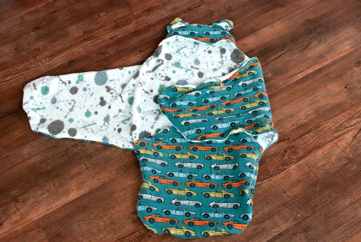 Genähter Wickelschlafsack mit blau-grünem Auto‑Muster und hellem, gesprenkeltem Innenfutter auf Holzboden