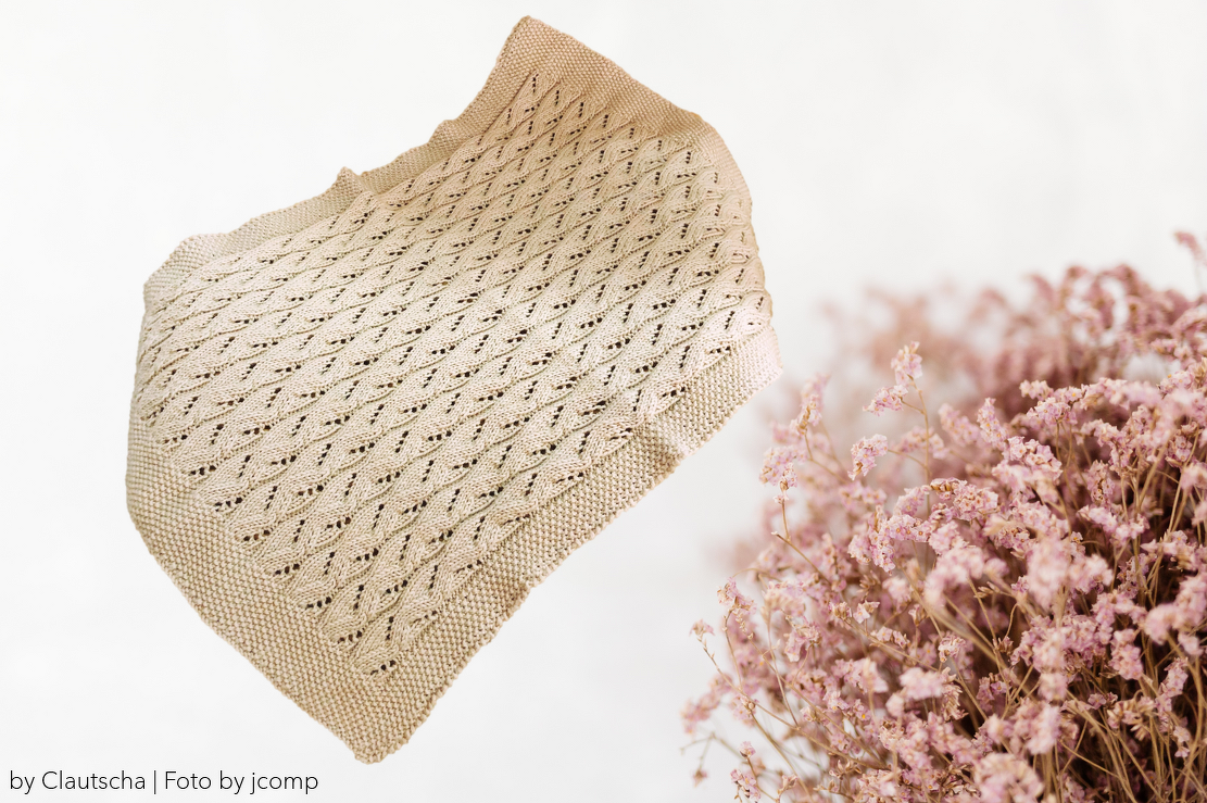 Hellbeige gestrickte Babydecke mit Rauten‑Lochmuster und kraus‑rechts gestricktem Rand auf hellem Hintergrund, daneben rosa Trockenblumen.