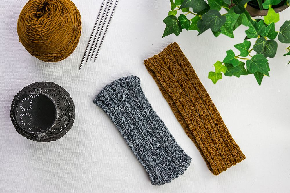 Zwei gestrickte Stirnbänder in Grau und Rostbraun mit Wollknäuel, Stricknadeln und einer Pflanze auf hellem Hintergrund.