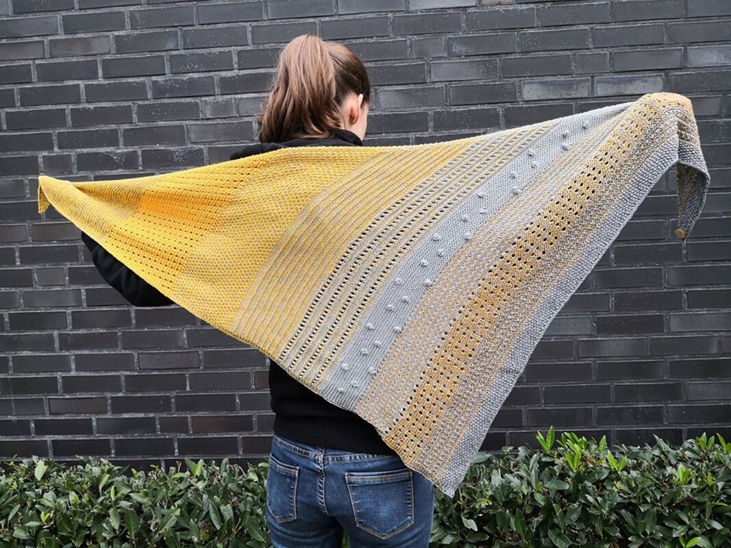 R
ckansicht einer Person, die ein gro
es gestricktes Dreieckstuch in Gelb und Grau mit strukturierten Streifen vor einer dunklen Ziegelwand h
lt.