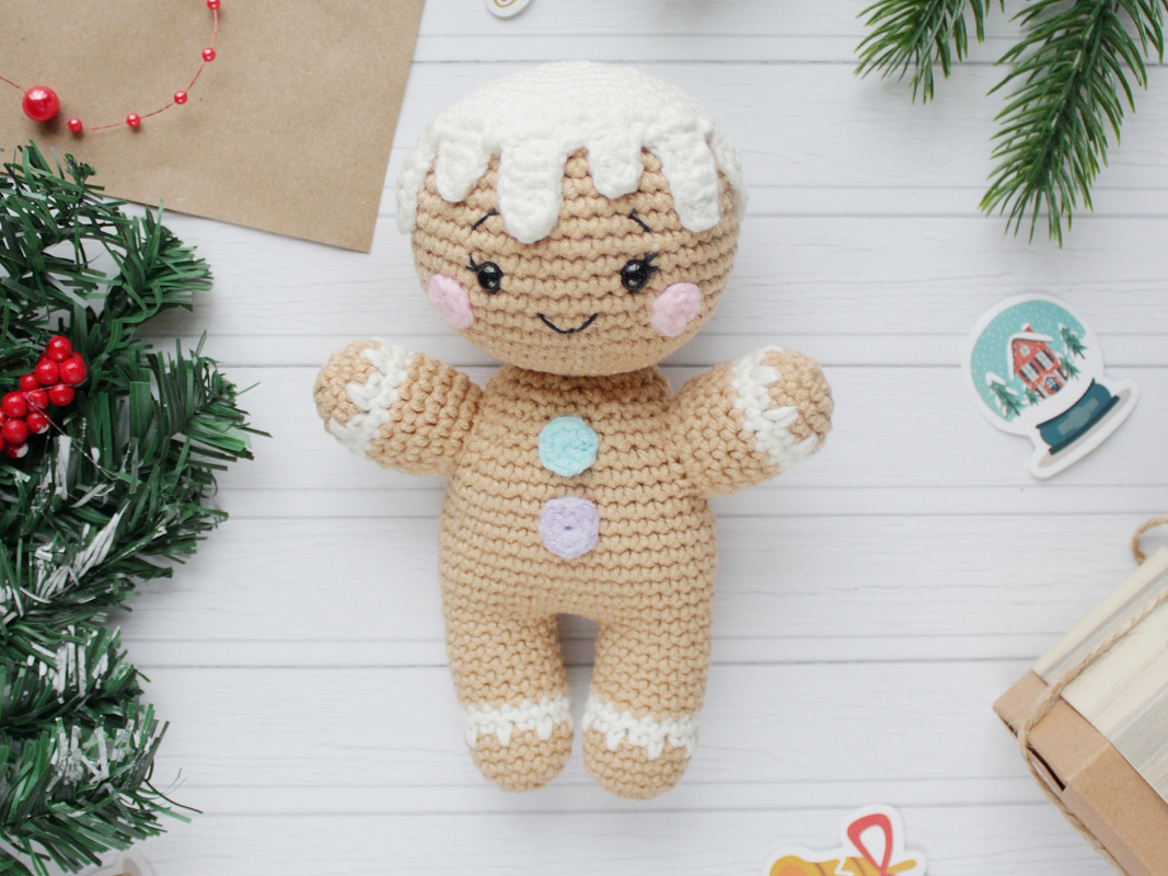 Gingerbread amigurumi doll with white icing trim, pastel buttons and a stitched smile on white wooden boards with evergreen decorations