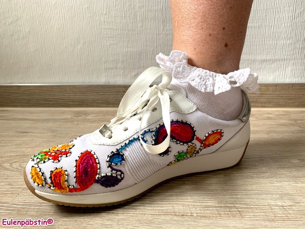 Nahaufnahme eines weißen Sneakers mit bunten textilen Verzierungen und einer weißen Socke mit gehäkeltem Rüschenabschluss.