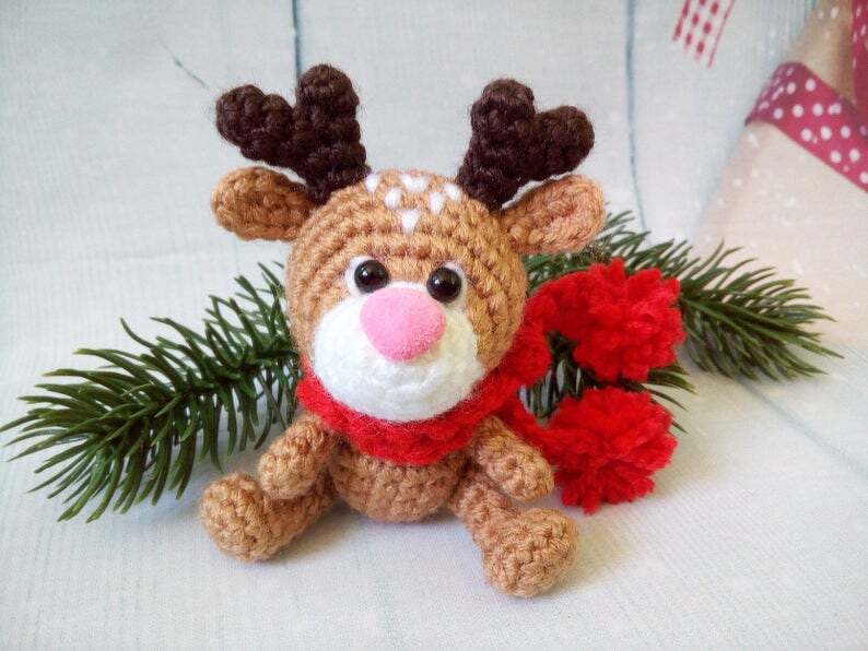 Small brown crochet reindeer amigurumi with red pom-pom scarf and pine sprig