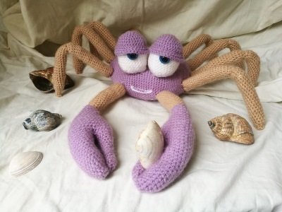 Gehäkelte lila-beige Krabbe mit großen Augen und Muscheln auf hellem Stoff