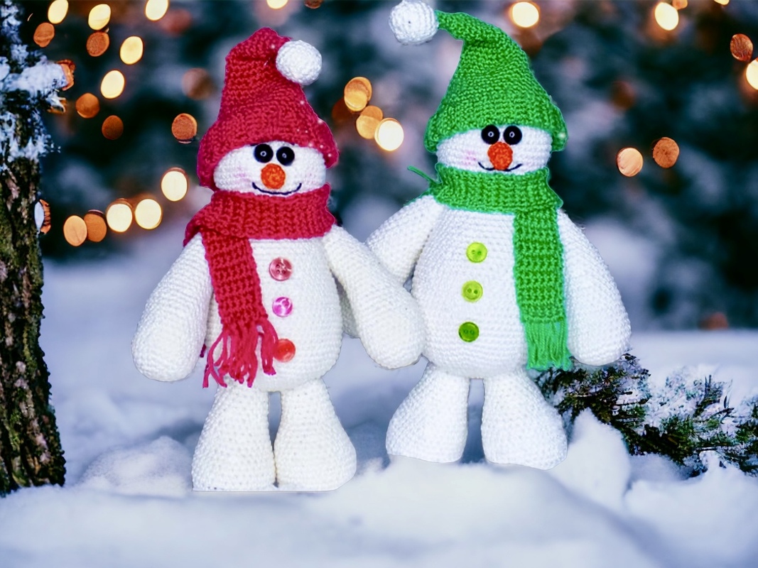 Two crocheted amigurumi snowmen in red and green hats standing on snow with blurred warm lights behind them