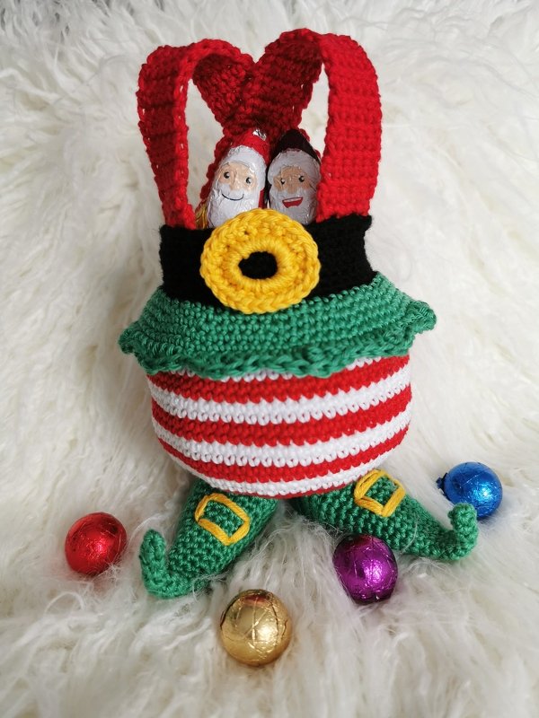 Gehäkelter Weihnachtskorb im Wichtel-Design mit roten Trägern, rot-weiß gestreiftem Körper, grüner Rüsche und Zipfel-Schuhen, gefüllt mit Schokoweihnachtsmännern.