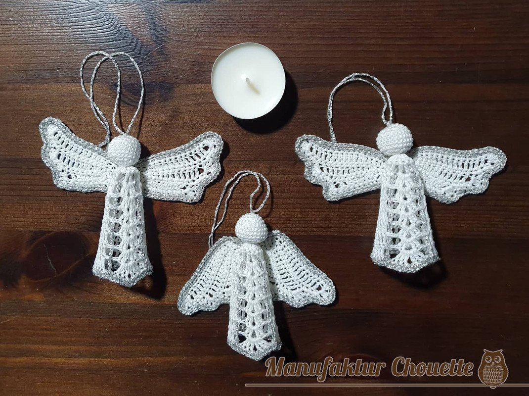 Drei weiße gehäkelte Engel mit silbernem Rand und Aufhängeschlaufen auf einem dunklen Holztisch neben einer Teelichtkerze.
