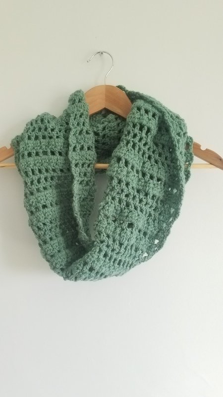 Sage-green crocheted infinity scarf with lacy openwork stitch hung on a wooden hanger