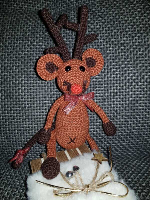 Braunes gehäkeltes Amigurumi-Mäuschen mit Rentiergeweih, roter Nase und Schleife, sitzend auf weißer Weihnachtsdeko vor grauem Hintergrund.