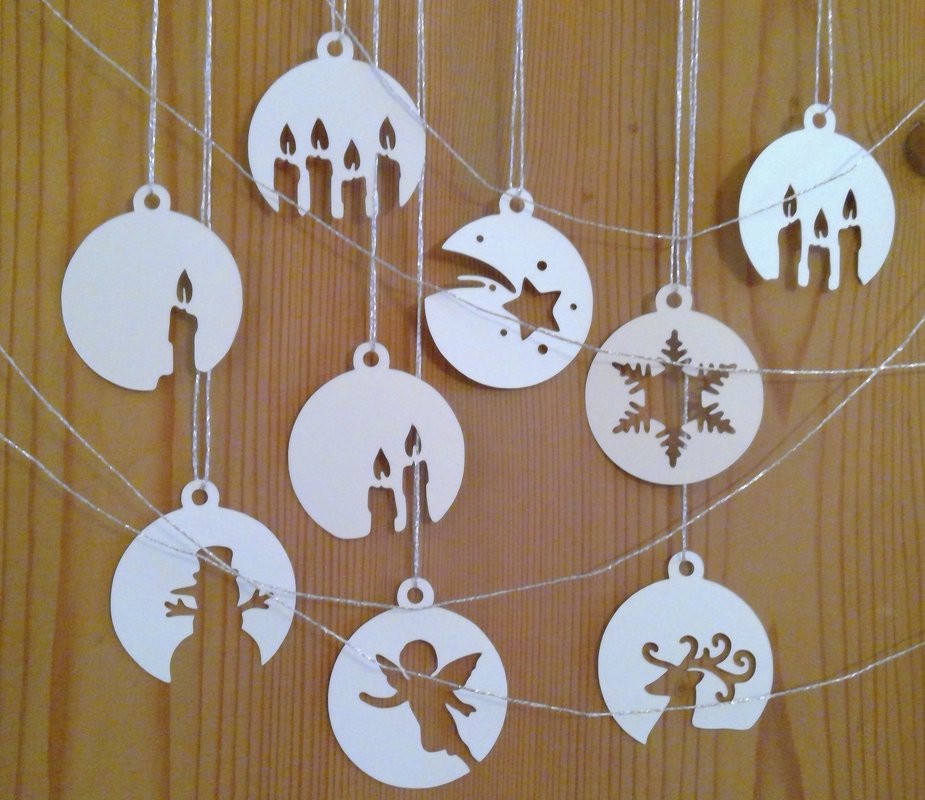 Weiße runde Weihnachtsanhänger mit ausgeschnittenen Silhouetten (Kerzen, Engel, Schneeflocke, Mond, Rentier, Schneemann) an silbernen Schnüren vor Holz