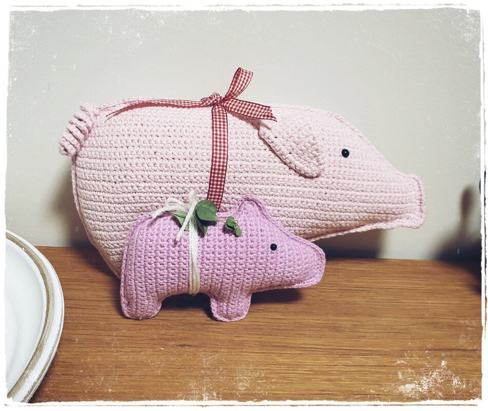 Zwei gehäkelte Schweine, ein großes rosa und ein kleines lila, auf einer Holzfläche.