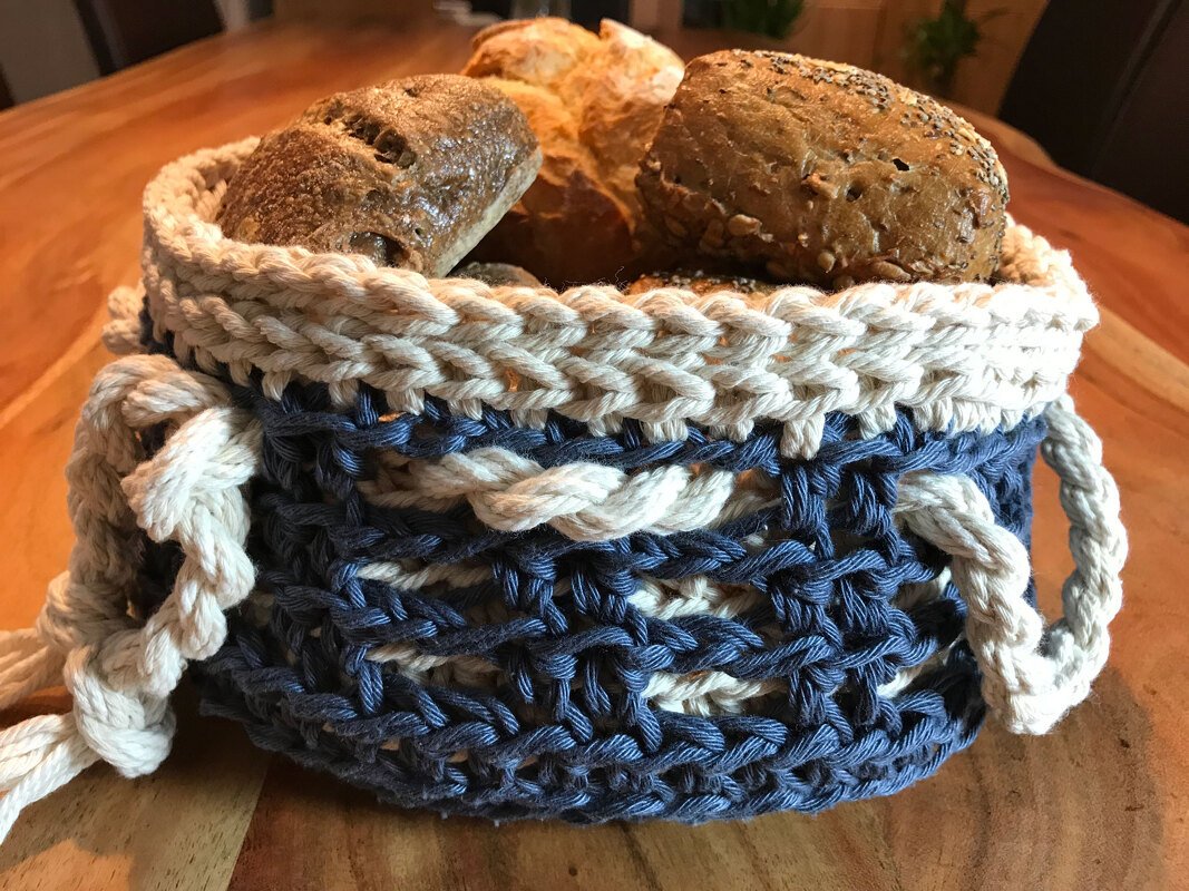 Zweifarbiger gehäkelter Korb aus dicker Kordel in Blau und Beige, gefüllt mit Brötchen auf einem Holztisch.