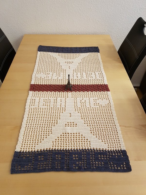Gehäkelter Tischläufer mit Eiffelturm- und Paris-Motiv in Cremeweiß, Dunkelblau und Rotbraun auf einem Holztisch.