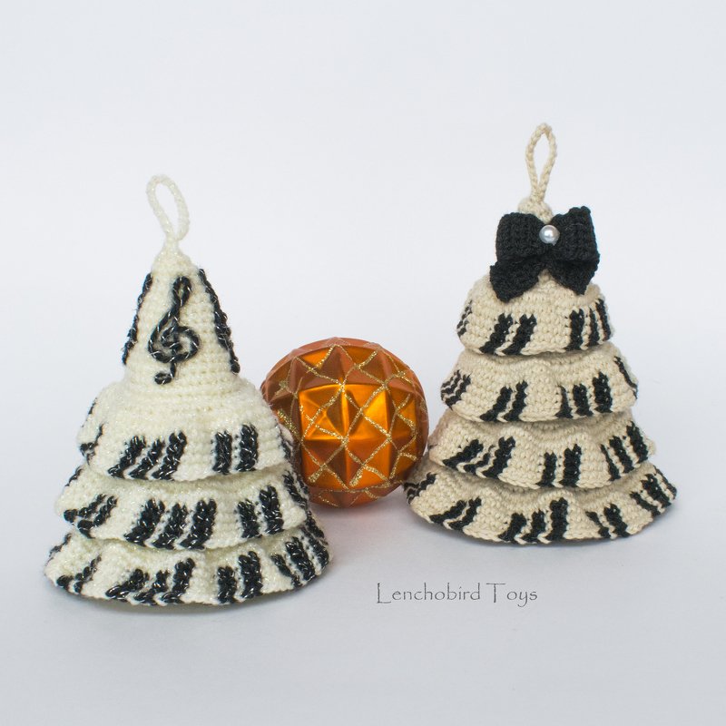 Two cream crochet tiered Christmas tree ornaments with black piano-key stripes and a treble clef detail beside an orange faceted bauble
